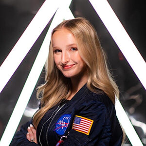 female with long blond hair wearing a blue jacket with NASA and American flag patches