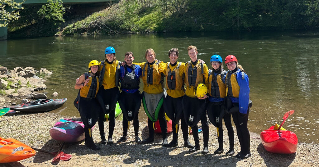 Students stand riverside in white water rafting gear at the Student Outdoor Leadership Experience.